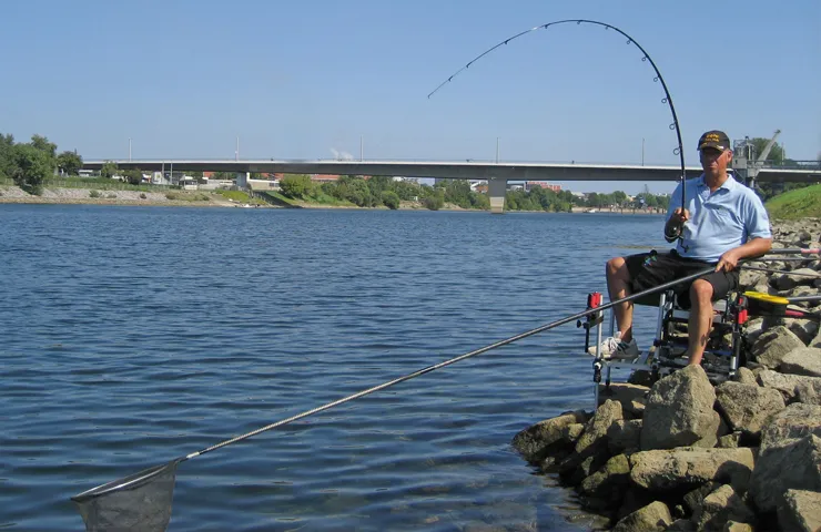 Angler sitzt an einem Fluss mit Angel und Kescher.